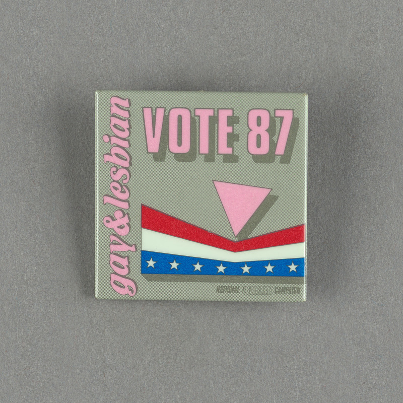 This square pinback button promoted the National Visibility Campaign for the Gay & Lesbian Vote. The pin reads “Gay & Lesbian / Vote 87” in pink and features a pink triangle seemingly pinching a distorted red, white, and blue starred banner. 