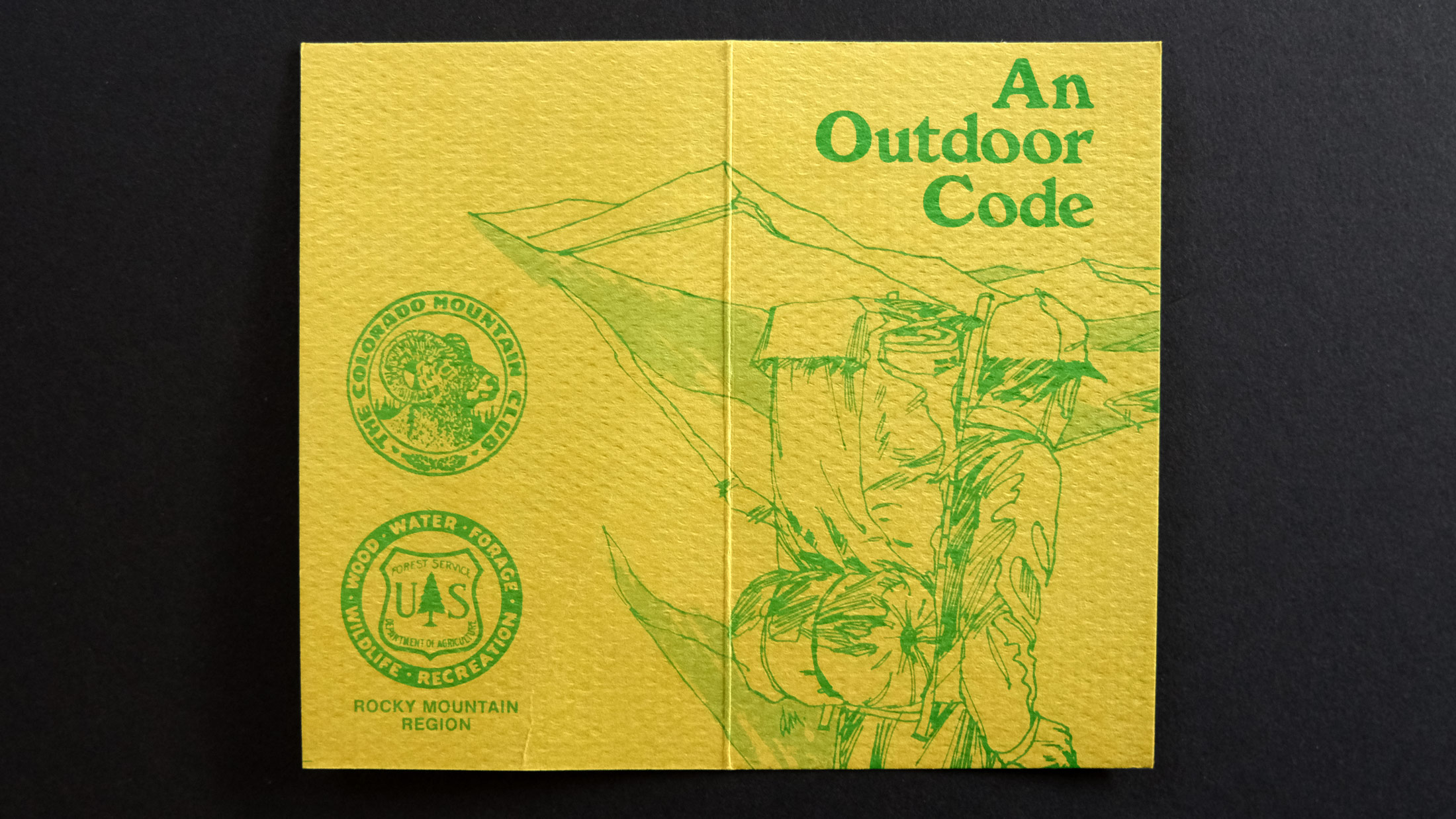 A small yellow pamphlet for the Forest Service has a green illustration of a backpacker in the mountains and it says An Outdoor Code in green serif font